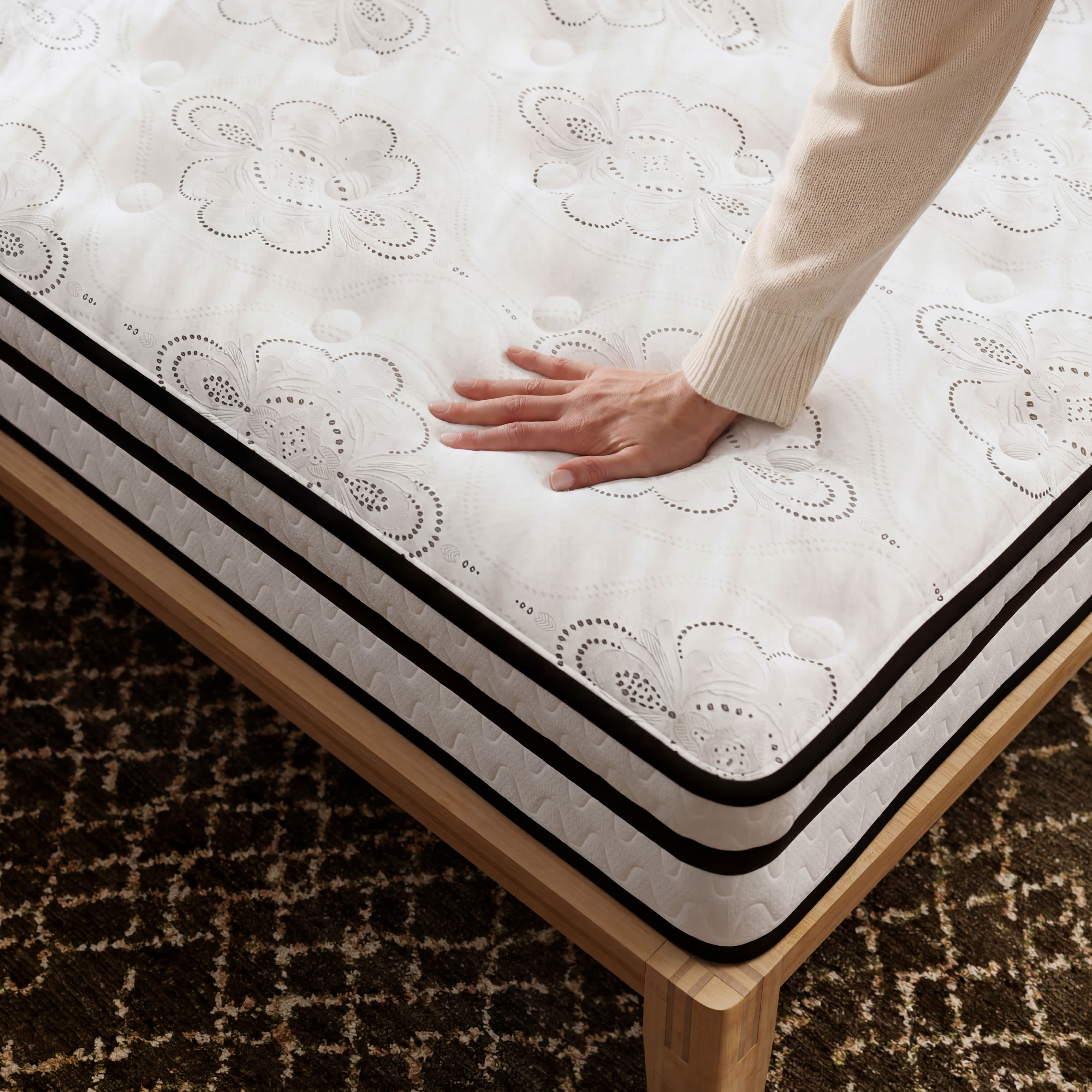 A hand presses into the white floral-patterned surface of a Hybrid mattress on a light wood bed frame