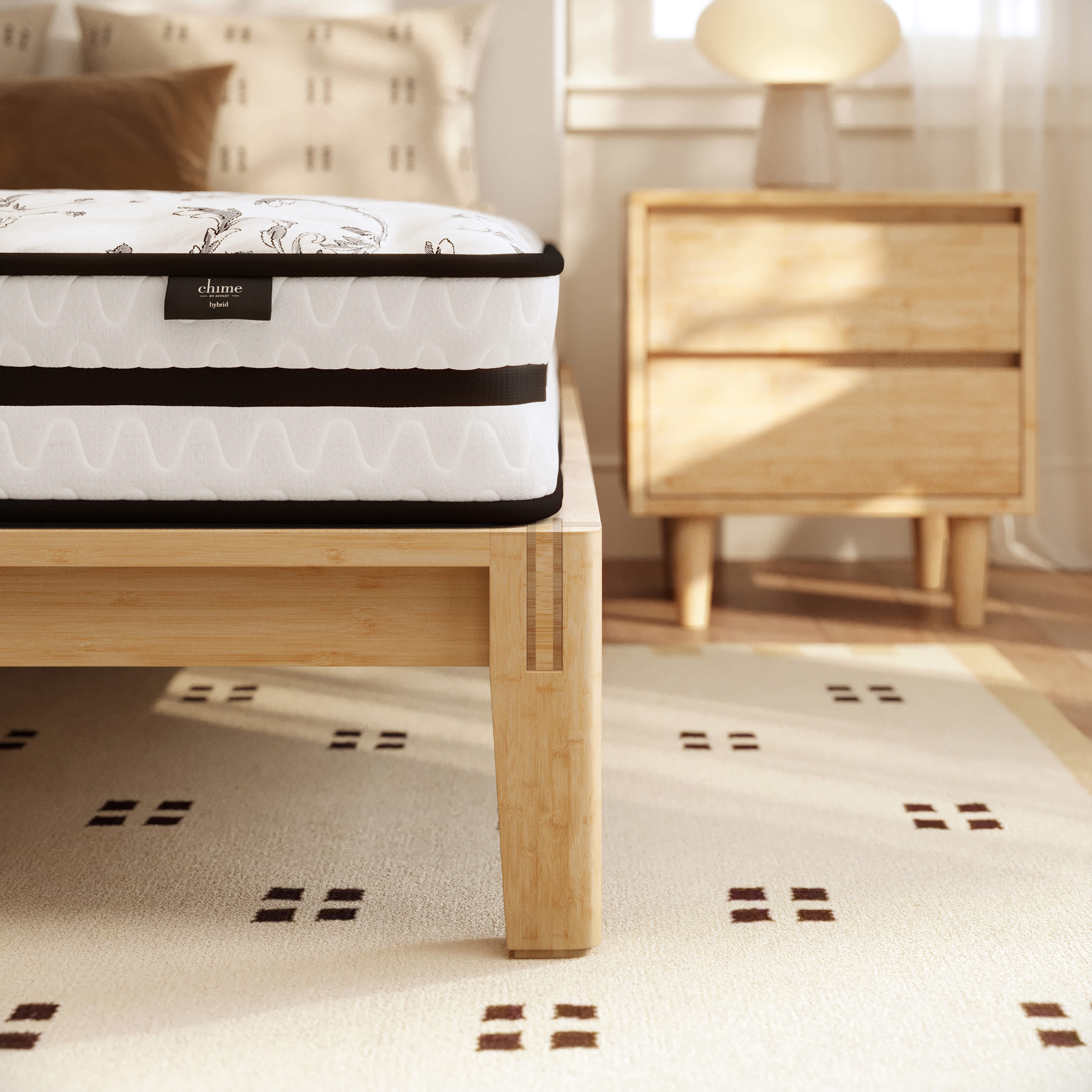 Hybrid mattress on a light wood bed frame in a bedroom with a patterned rug and nightstand