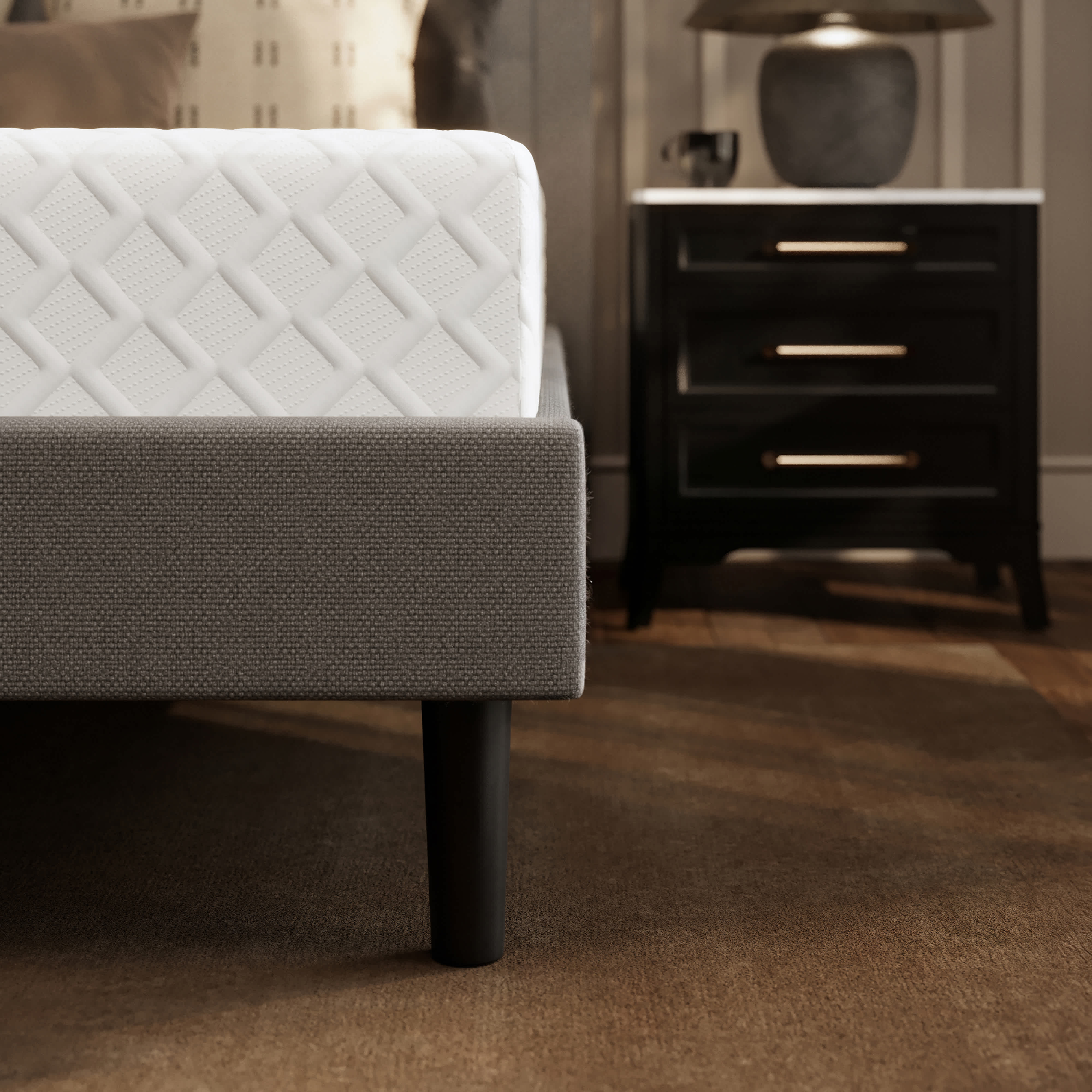 A Memory Foam mattress on a gray upholstered bed frame with black legs, next to a dark nightstand