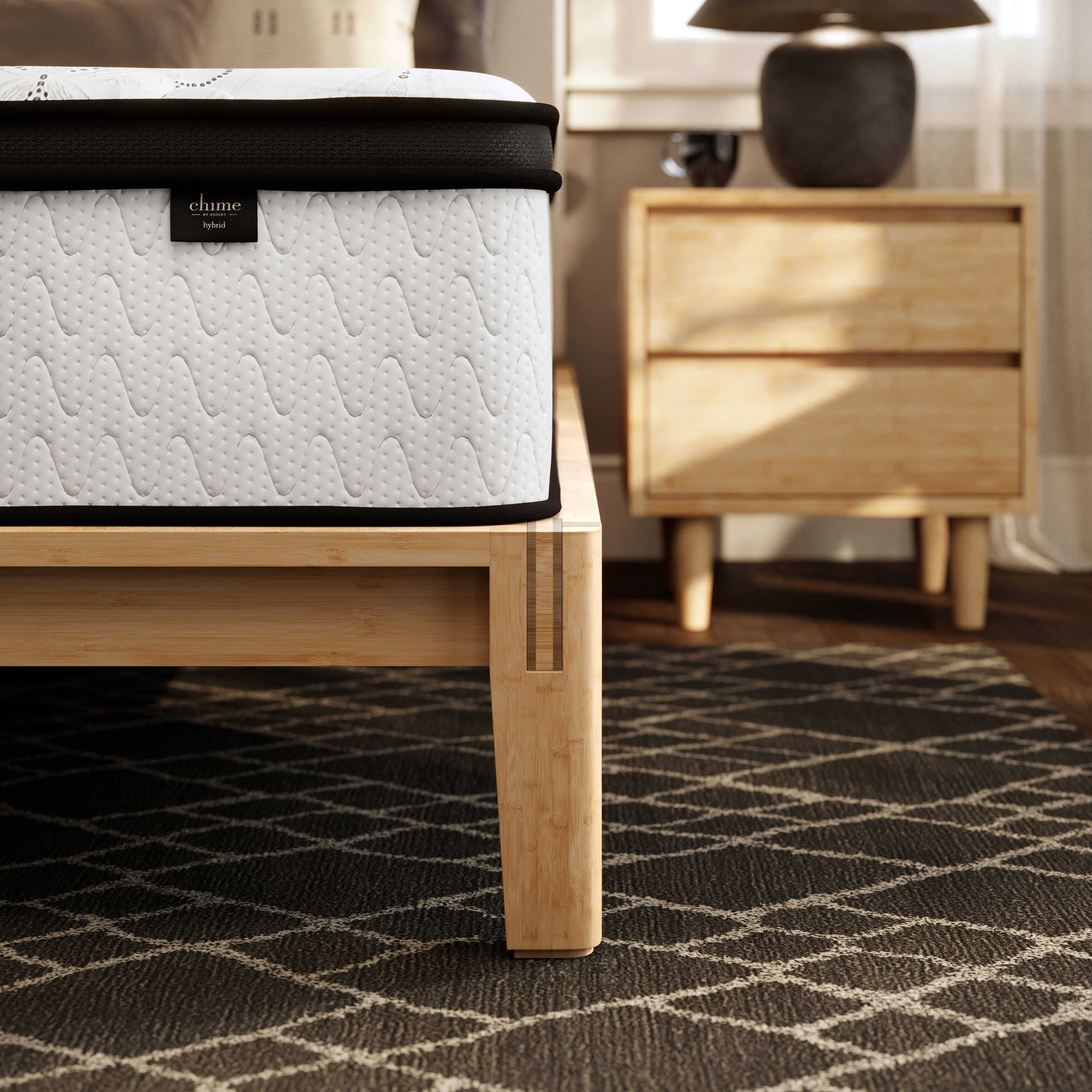 A Hybrid mattress on a light wood bed frame, with a nightstand and lamp in the background