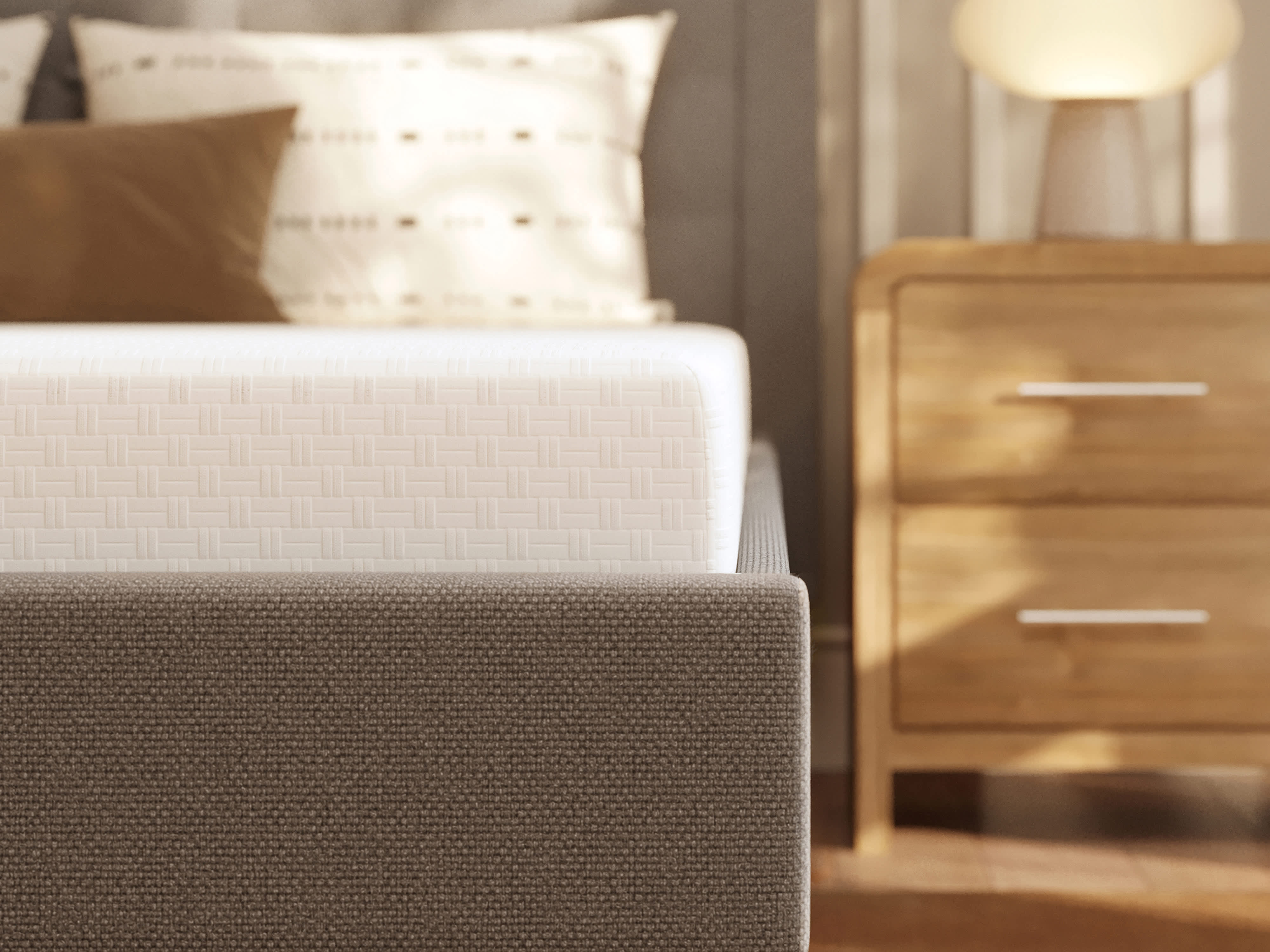 A close-up of a bed with a white memory foam mattress and a gray fabric frame, next to a wooden nightstand.