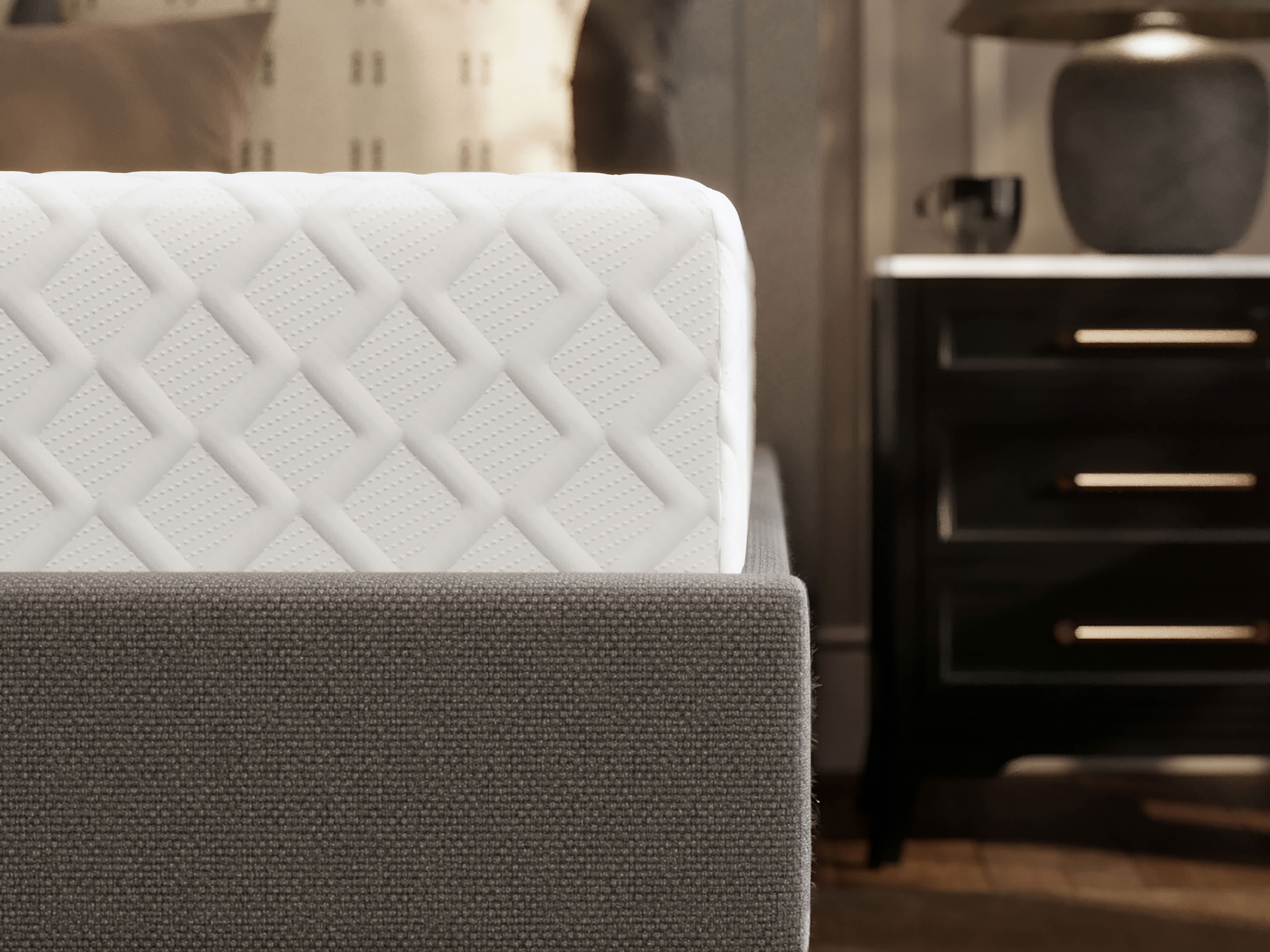 A memory foam mattress on a gray upholstered bed frame with black legs, next to a dark nightstand.