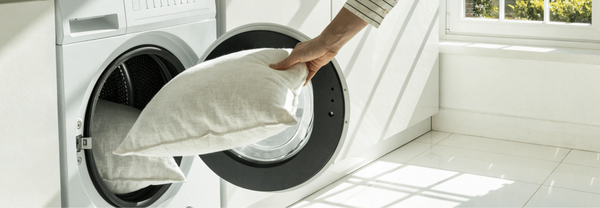 Two pillows going into a front-load washing machine, clean white laundry setting, bright and airy