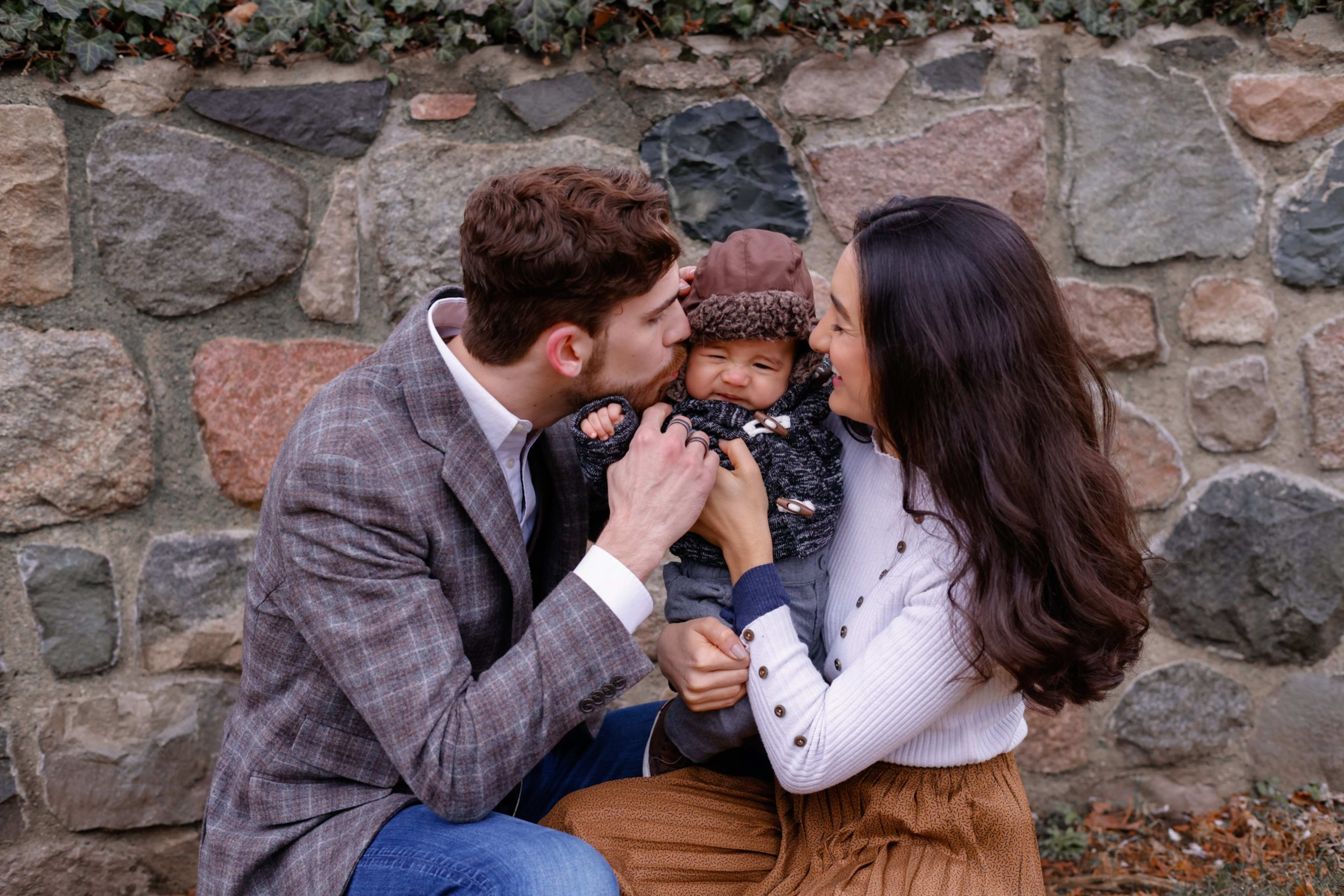 Parents kissing their baby by a stone wall in autumn, southeast Michigan family session