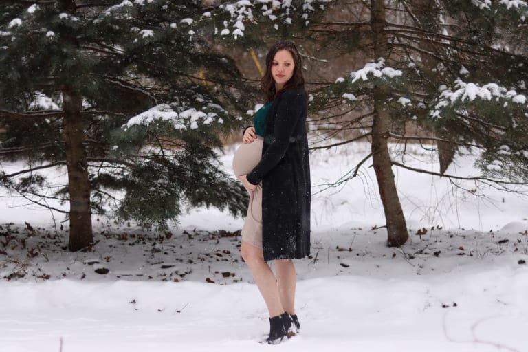 Mother-to-be cradling her bump in a snowy pine forest — a winter maternity session in Metro Detroit