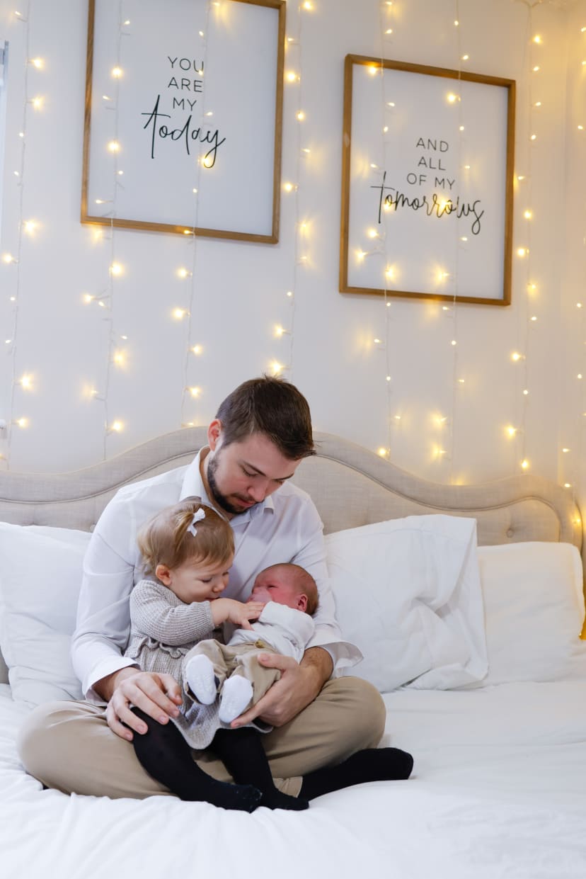 Father on a bed with his toddler and newborn under fairy lights, Metro Detroit newborn session