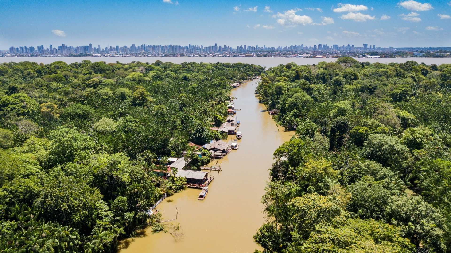 Aerial view of a narrow brown river cutting through dense green rainforest near Belém, Pará, Brazil. Small wooden houses and boats line the riverbanks. In the distance, across a wide bay, the modern skyline of Belém with high-rise buildings is visible under a bright blue sky