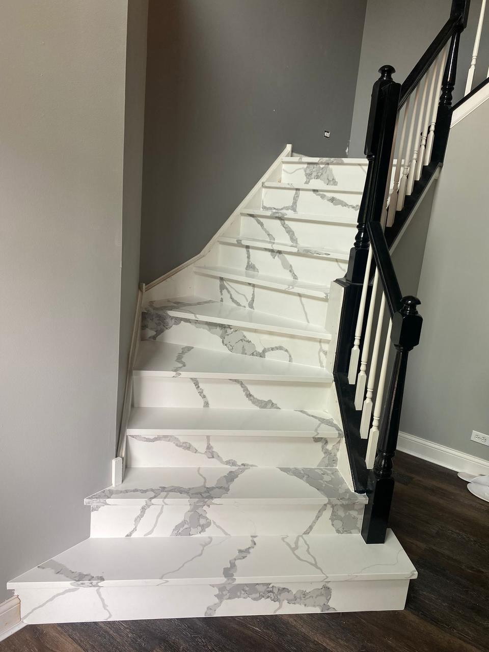 Stone staircase with white treads featuring gray veining, black railing, and a gray wall backdrop, showcasing custom design and craftsmanship for elegant indoor spaces.