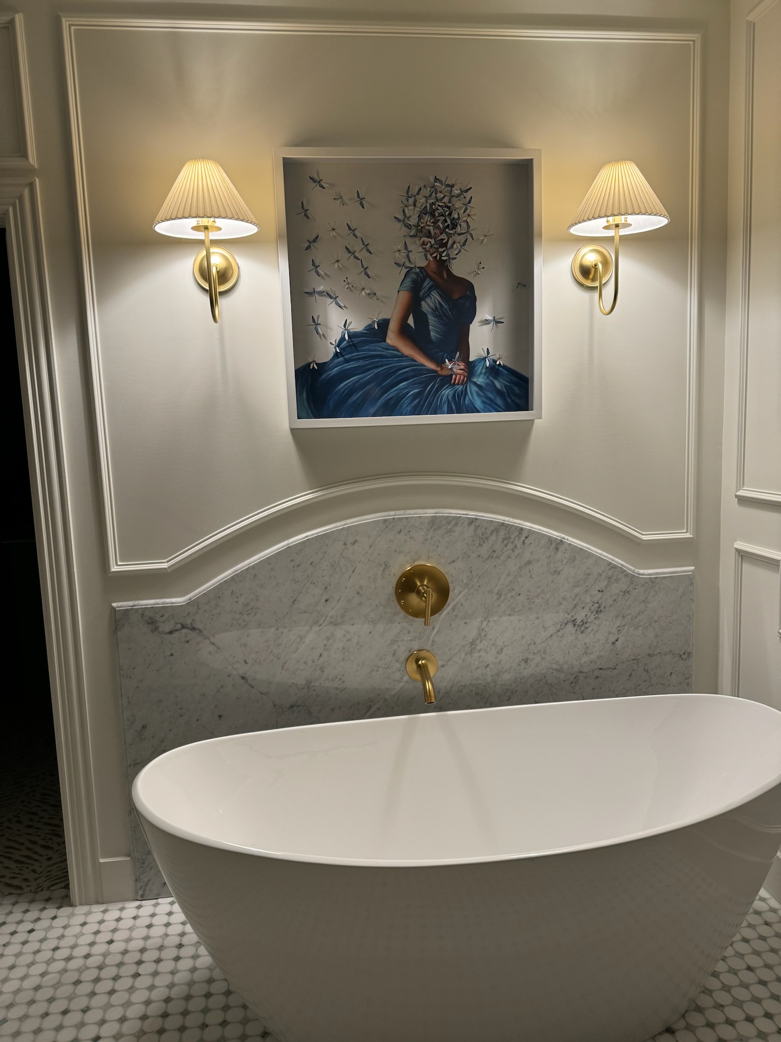 Elegant bathroom featuring a freestanding white bathtub, gold fixtures, and a striking artwork of a woman in a blue dress adorned with dragonflies, complemented by soft wall sconces and marble accents.