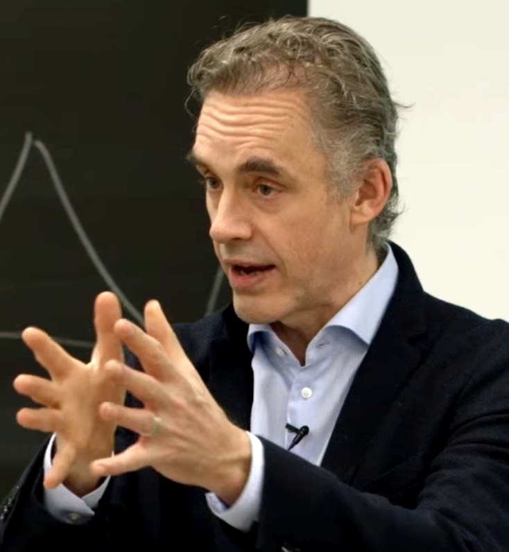 Jordan Peterson mid-gesture during a lecture, hands raised in front of a chalkboard