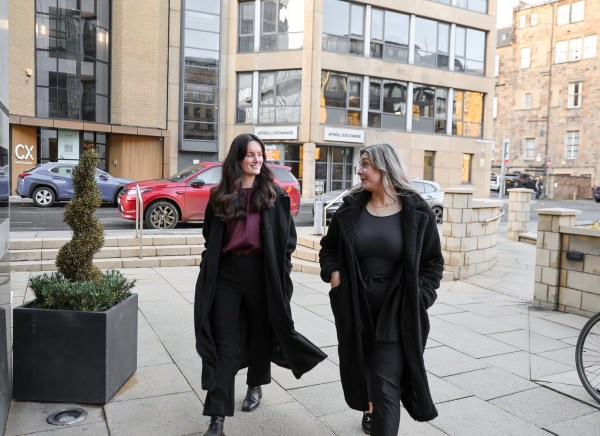 Two women walk together on a city sidewalk, smiling and chatting.