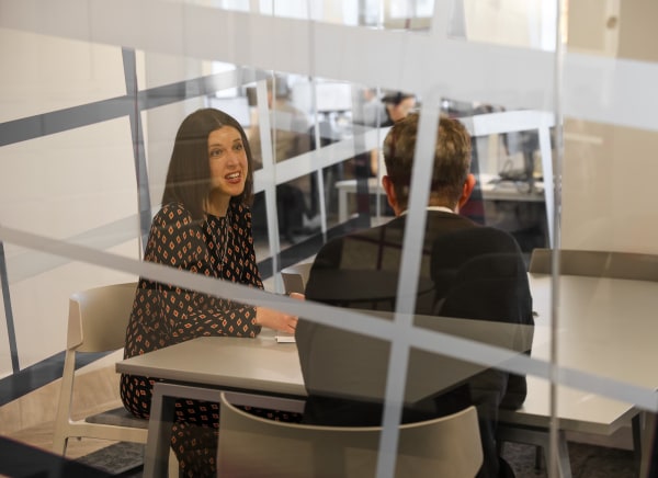 A woman talks animatedly to a man in a modern office setting, separated by glass panels.