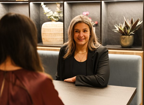 Two women converse at a table in a modern setting with decorative plants in the background.