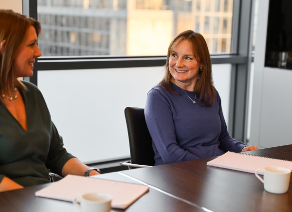 Two women smiling in a meeting room with notebooks and coffee cups on the table.