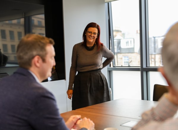 A woman stands at a conference table, engaging with seated colleagues in a modern office.
