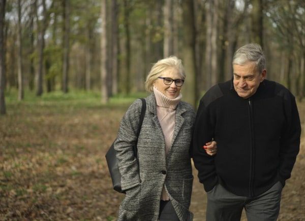 A happy elderly couple walks arm in arm through a wooded park.