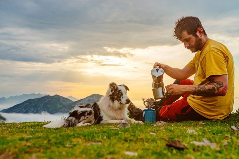 A man with his dog for a walk by the water||An Alsatian chasing a frisbee thrown by his owner in the forest||young mountaineer man has breakfast drinking coffee with his dog in the mountains
