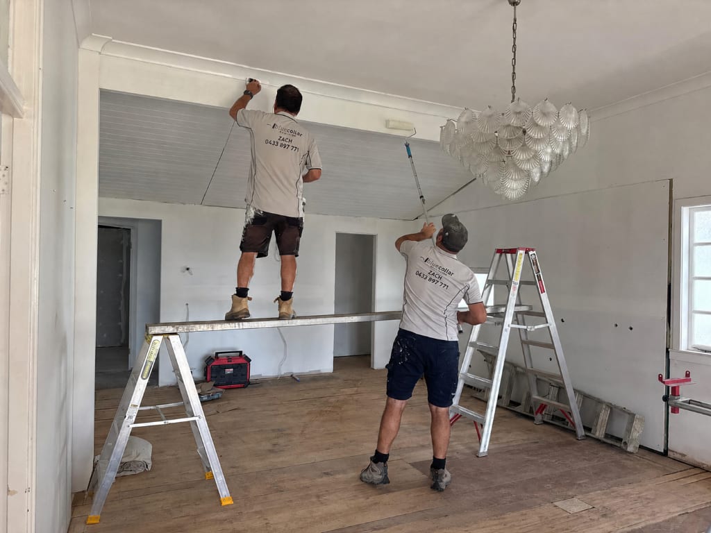 Two Blue Collar Painting tradespeople in branded uniforms painting the interior ceiling and walls of a classic Queenslander home on the Gold Coast