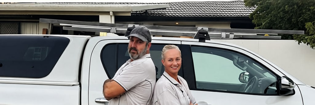 Zach Franses and his wife in Blue Collar Painting uniforms standing back to back in front of branded ute at sunset, Gold Coast