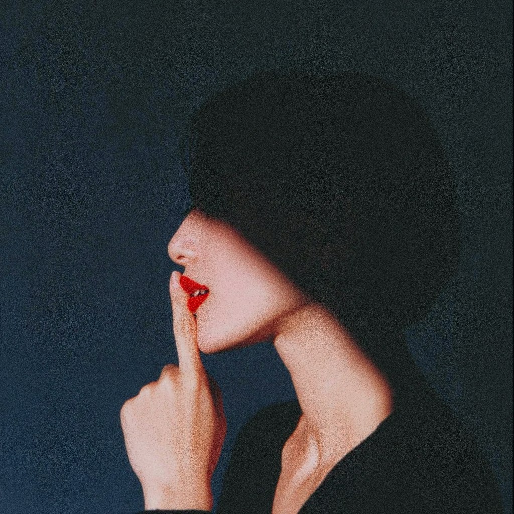 A person with red lipstick and a black hat, holding their finger to their lips.