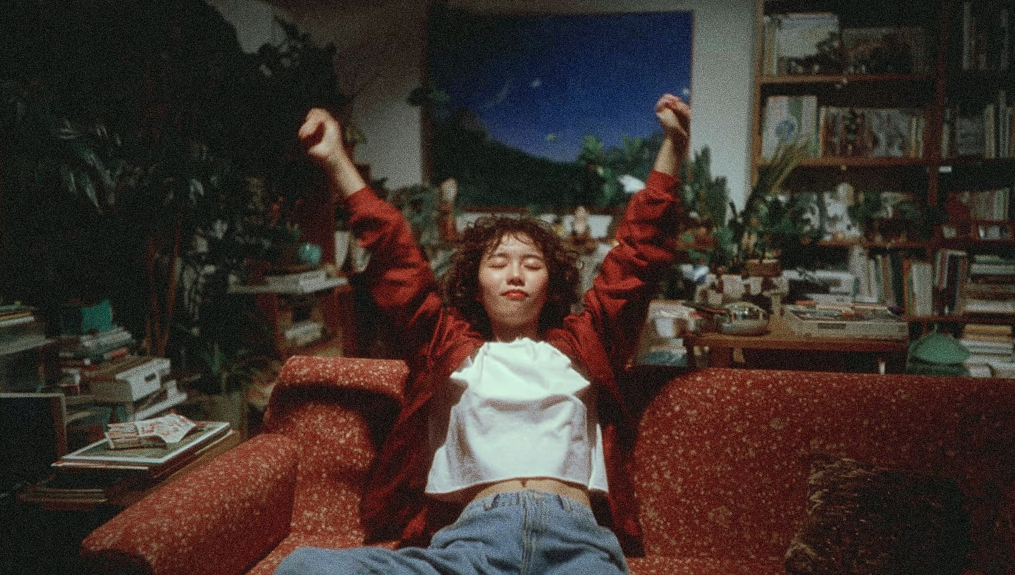 A person stretching with arms raised in a cozy room filled with books and plants.