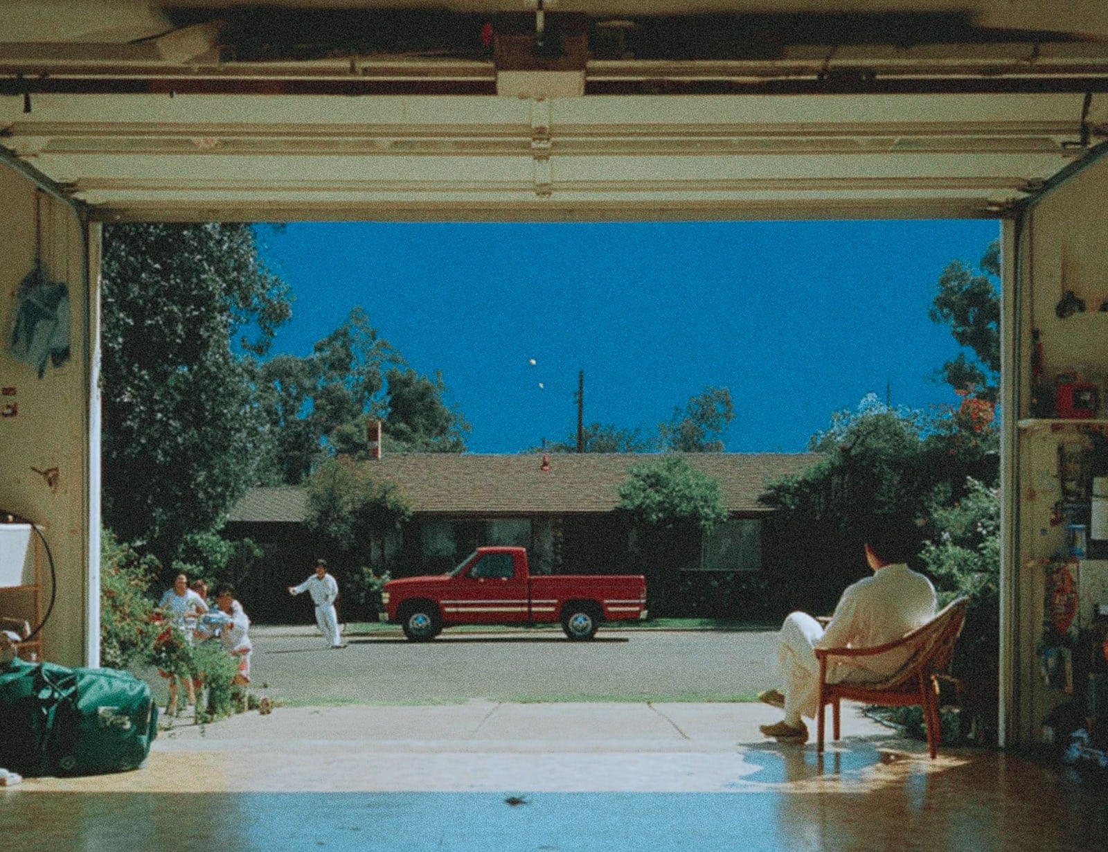 A person sitting in a chair in a garage with a red truck and a family outside.