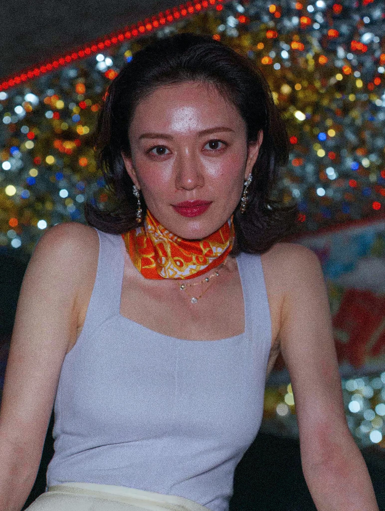 A person wearing a light grey sleeveless top and an orange scarf with a necklace and earrings.