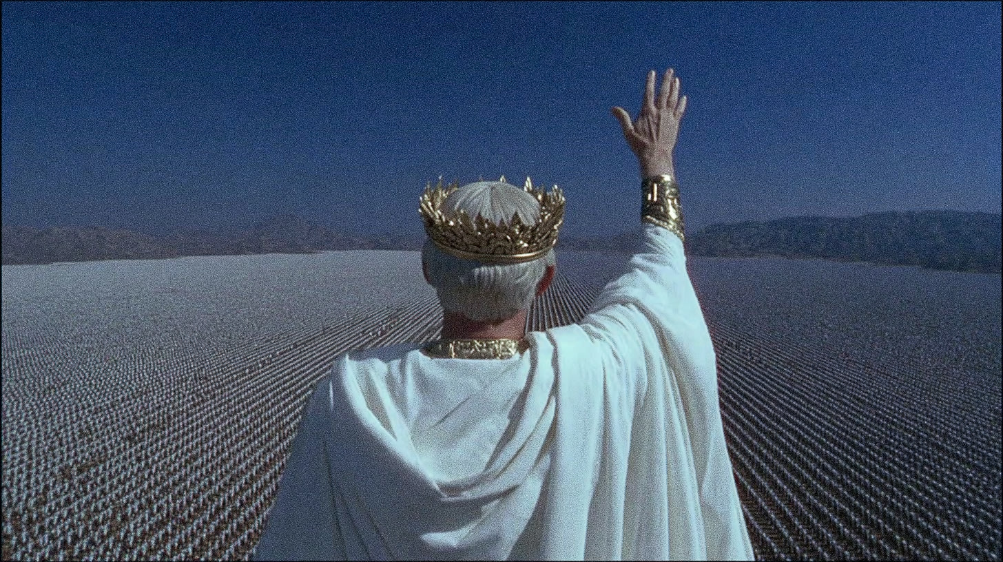 A person in a white robe and crown stands with one arm raised in a vast, flat landscape under a clear blue sky.
