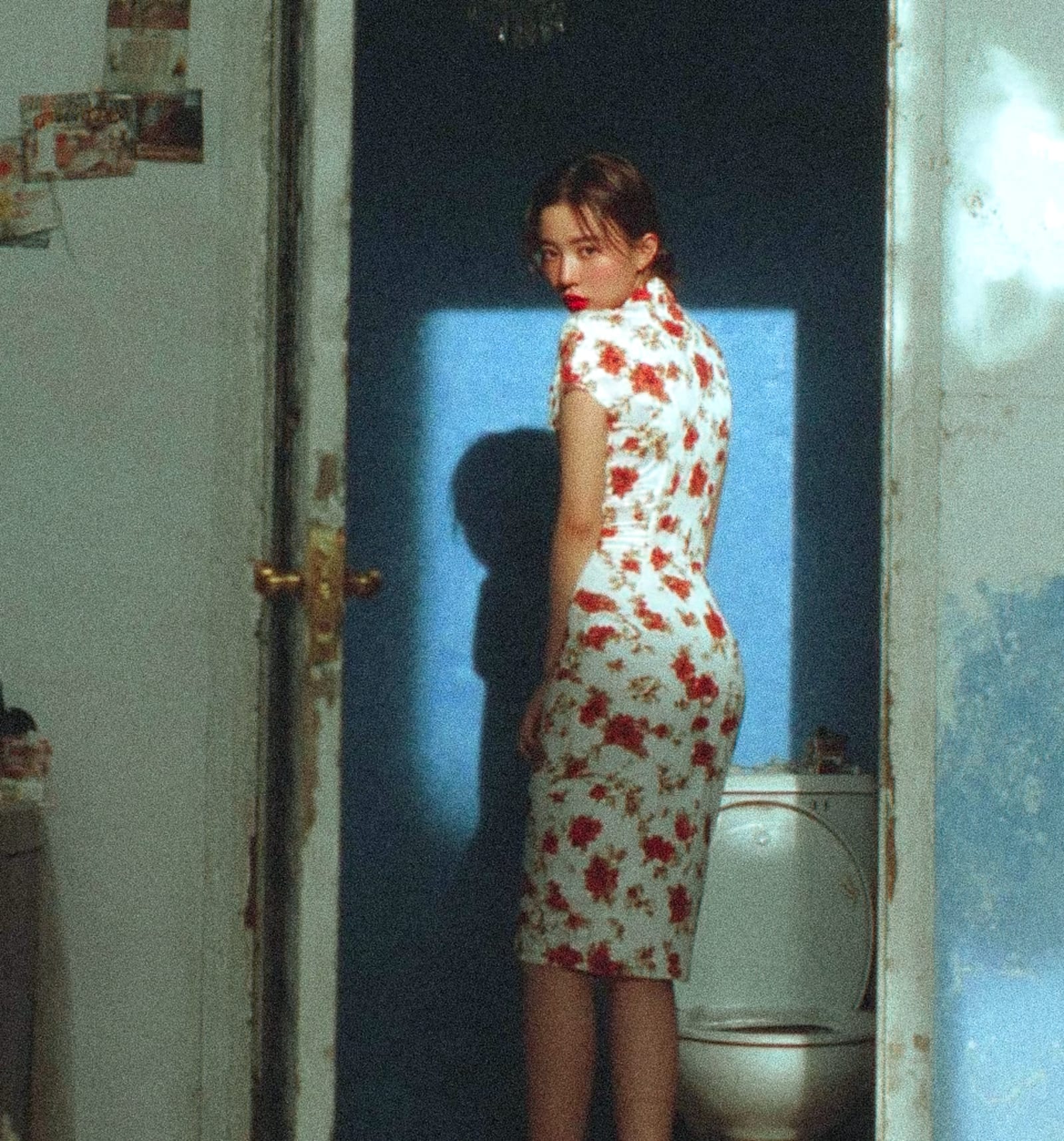 A person wearing a floral dress standing in a dimly lit room with a toilet in the background.