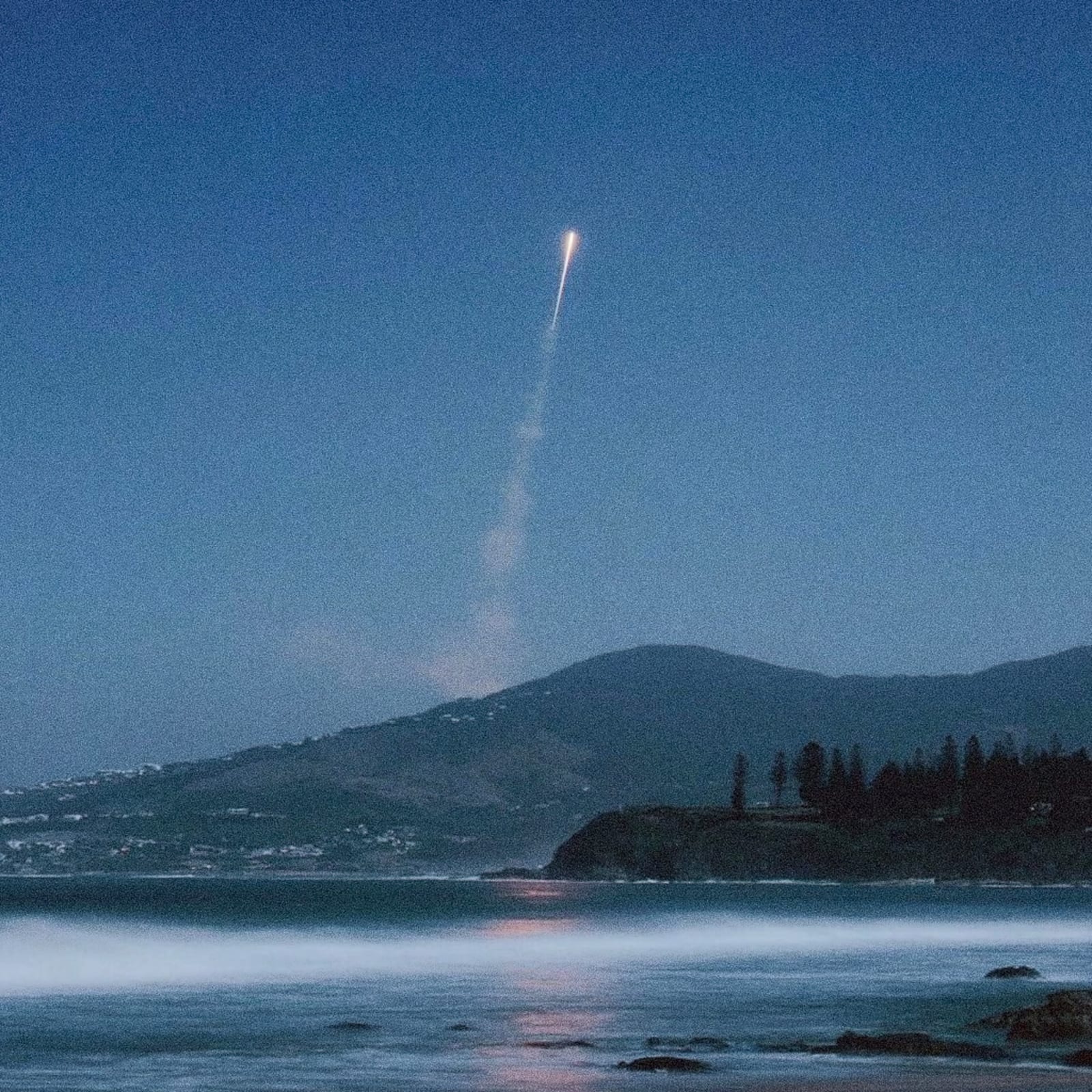 A rocket launches into the night sky from a coastal area with a mountainous background and a body of water in the foreground.