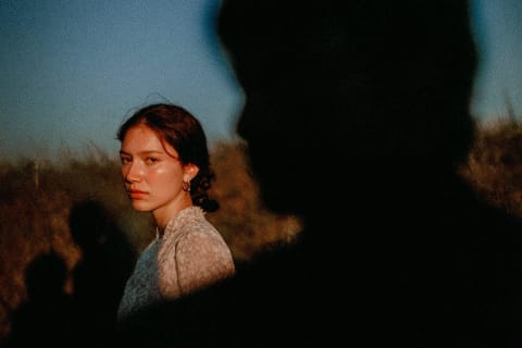 A person with dark hair and earrings standing in a field with a silhouette in the background.