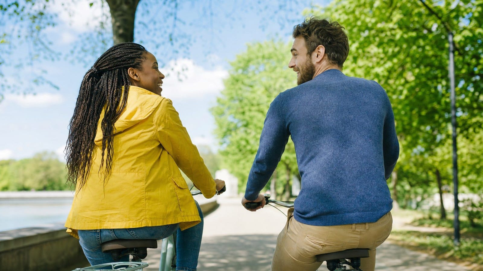 A couple rides bicycles together on a tree-lined park path.