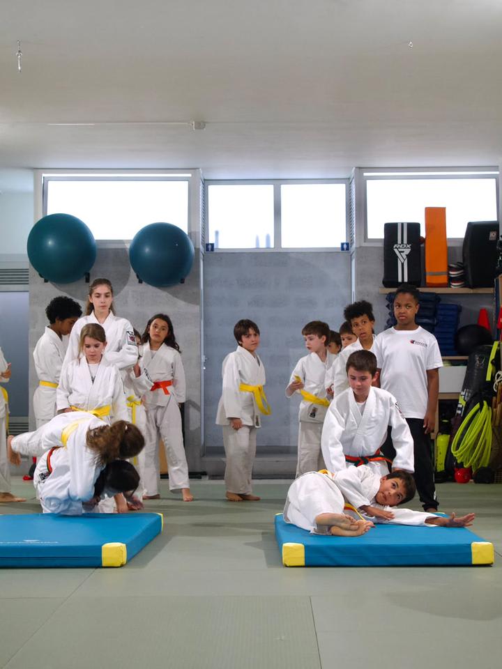 Crianças de quimono a aprender a cair em colchões, observadas pela turma