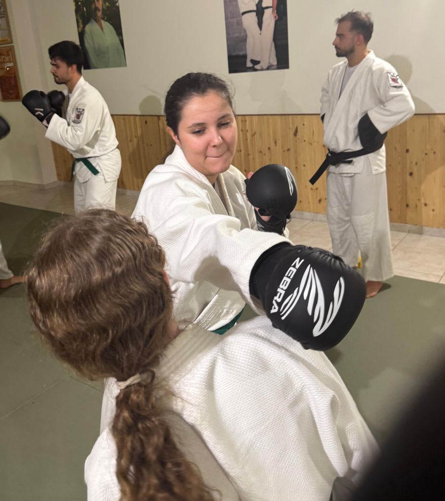 Aluna a sorrir enquanto bate nas luvas de um colega numa aula de Krav Maga, com outros alunos a treinar atrás