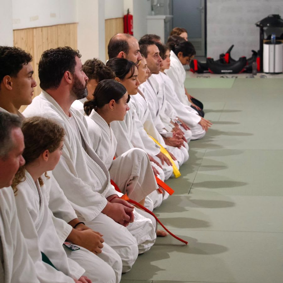 Alunos adultos de quimono sentados em linha no tatami, no início de uma aula de Krav Maga