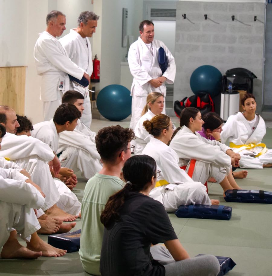 Alunos de várias idades sentados no tatami a ouvir três instrutores no início de uma aula de Krav Maga