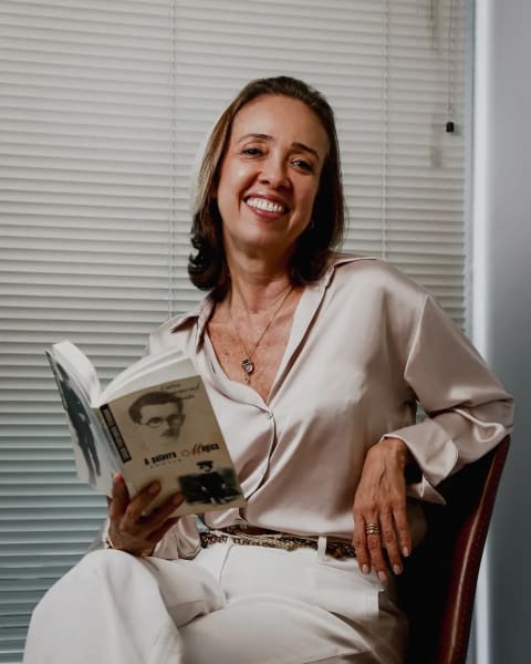 Jaq Priston sentada em uma poltrona, com um livro nas mãos, em seu ambiente de trabalho.