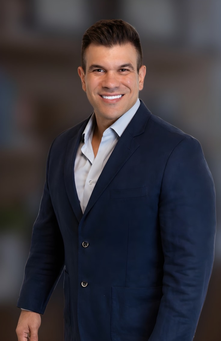 Dr. Rodrigo Schröder sorrindo no consultório, de blazer azul-marinho e camisa azul-clara