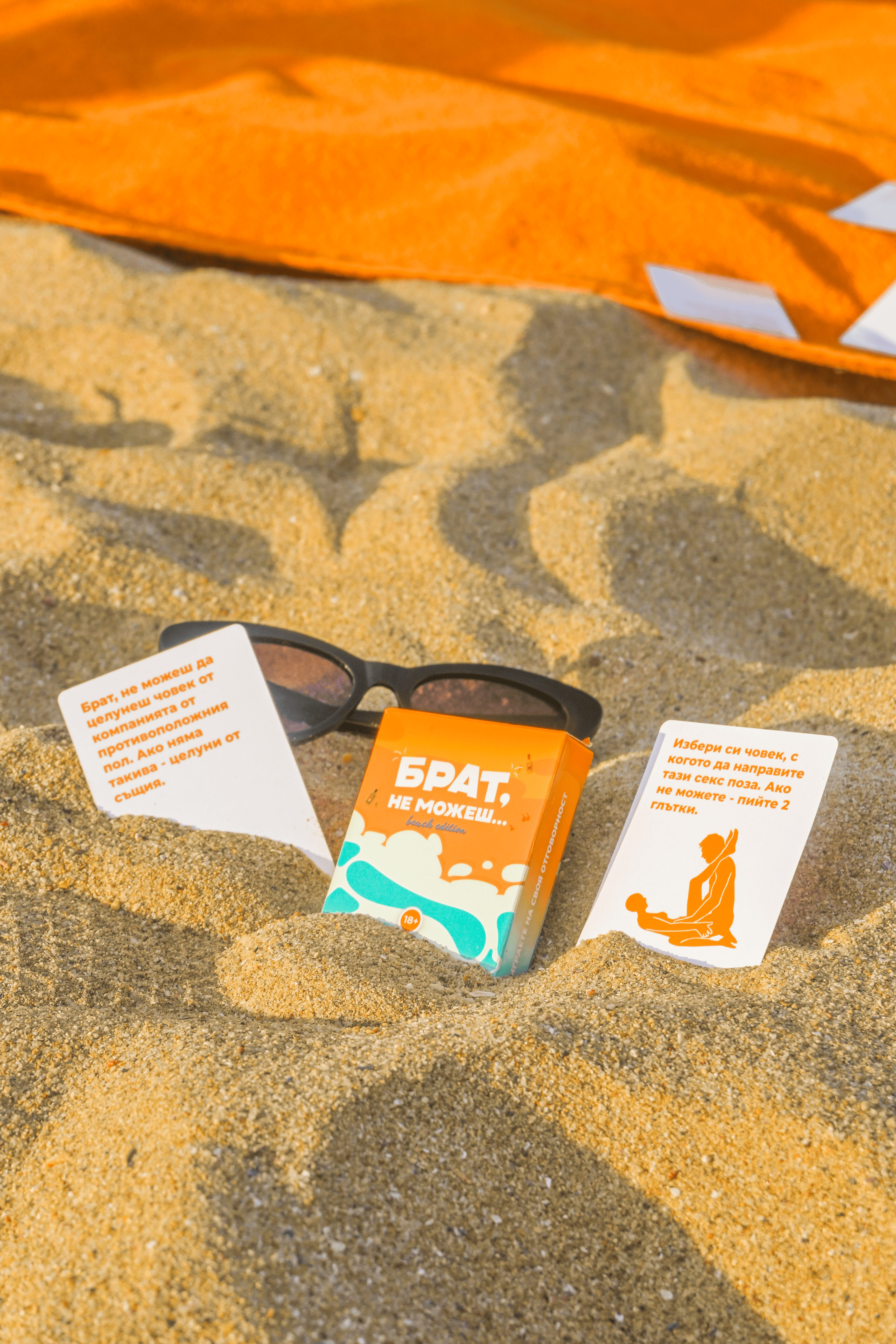 Кутия на парти игра Брат, не можеш – Beach Edition, поставена в пясъка между две предизвикателства и слънчеви очила