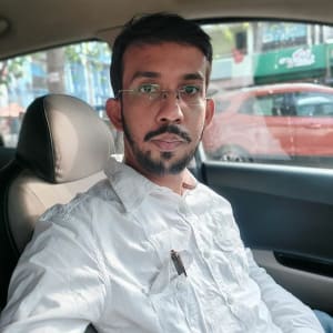 DG10 - A man with a beard and glasses sits inside a car. He is wearing a white collared shirt. The car's window shows a red vehicle and blurred street scenery outside. Light filters in through the window, creating a natural, daytime ambiance.
