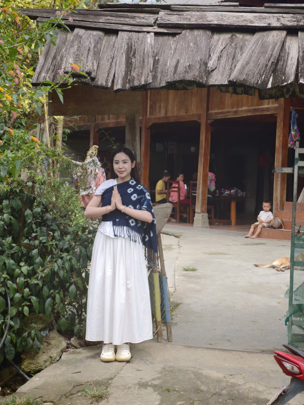 A young woman wearing an indigo-dyed scarf in front of a wooden-shingled stilt house in Ít Thái village