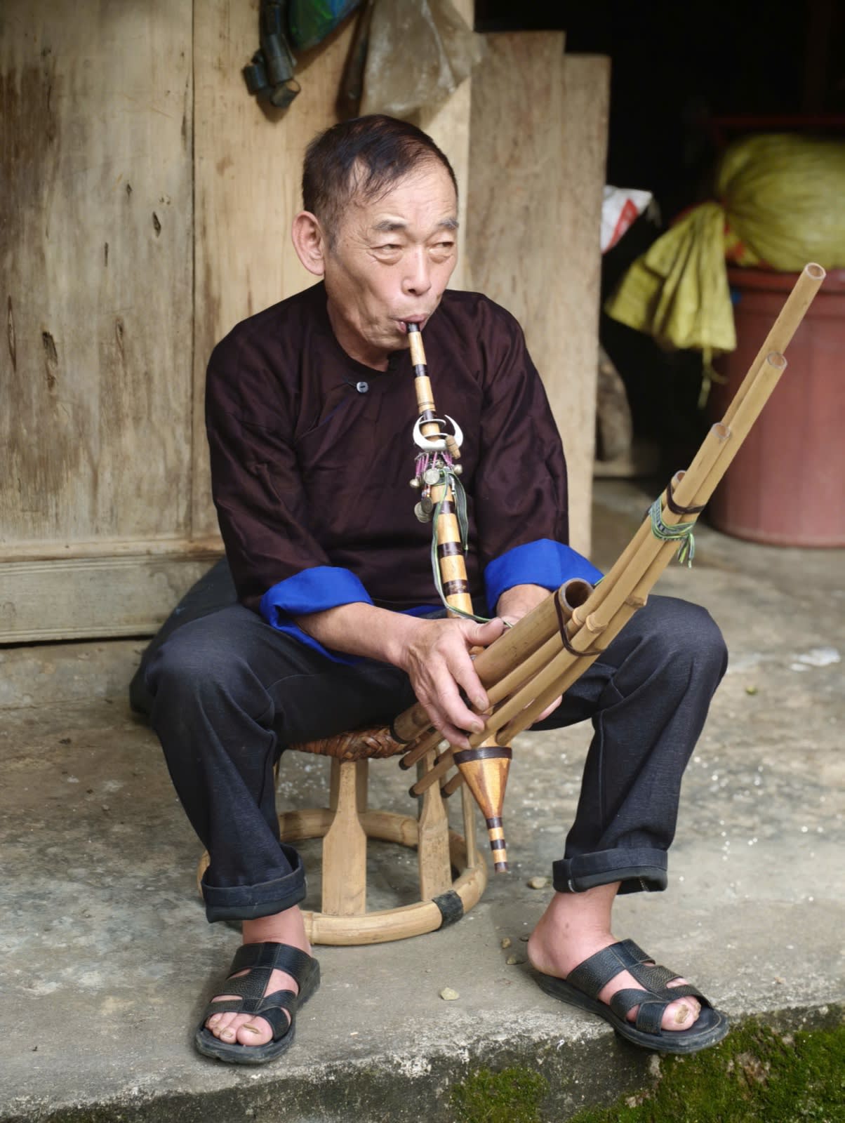 An elderly Hmong man playing the khèn on the porch of a wooden house in Lìm Mông village