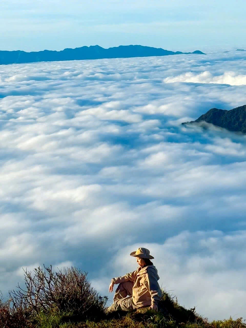 A trekker sitting on the summit of Lùng Cúng, looking down on a sea of clouds