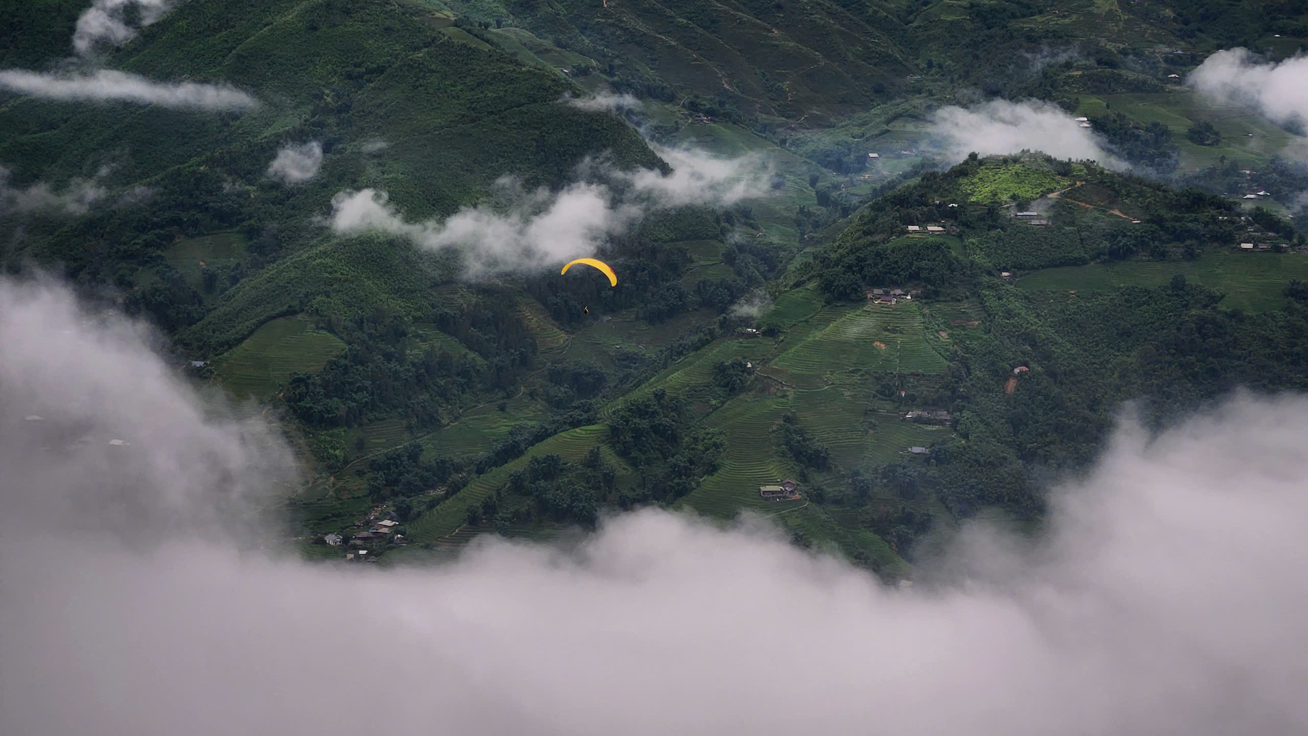 A yellow paraglider flying between low cloud banks over the Sapa hills