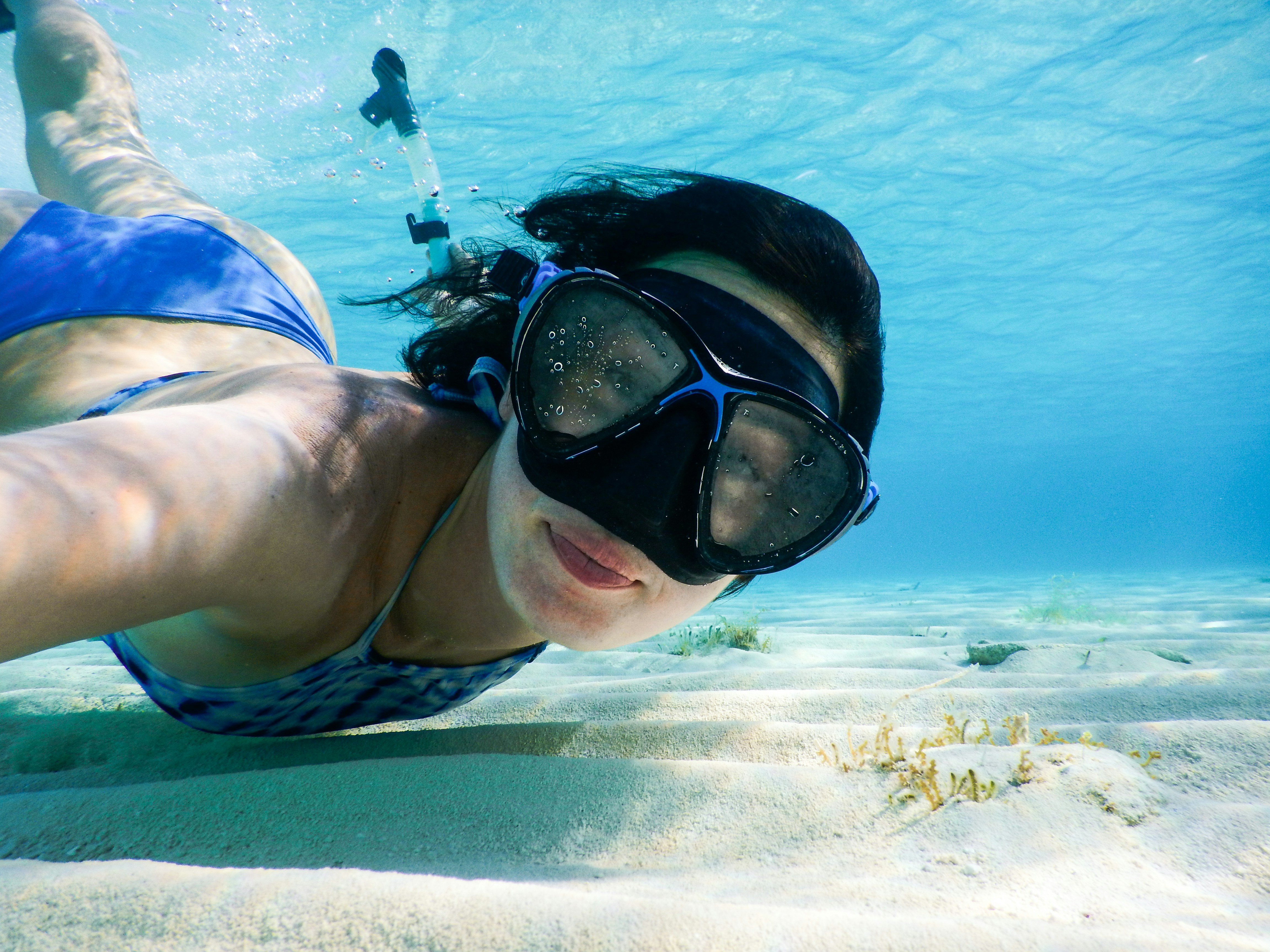 A person snorkeling underwater, wearing a mask and swimming over the sandy seabed with clear, blue water.