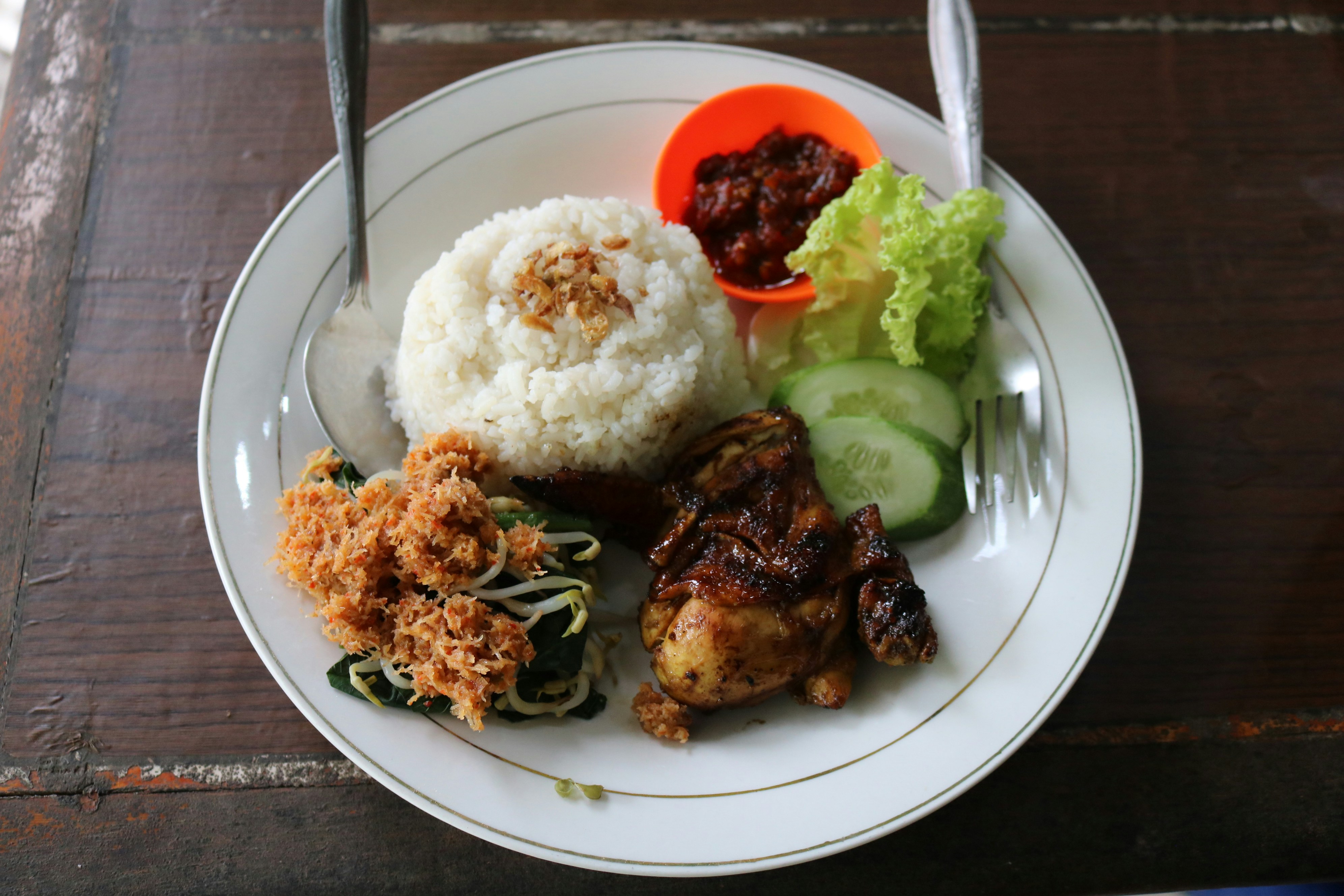 A plate of Indonesian nasi campur, featuring rice, grilled chicken, sautéed vegetables, crispy shallots, and a side of sambal and cucumber.