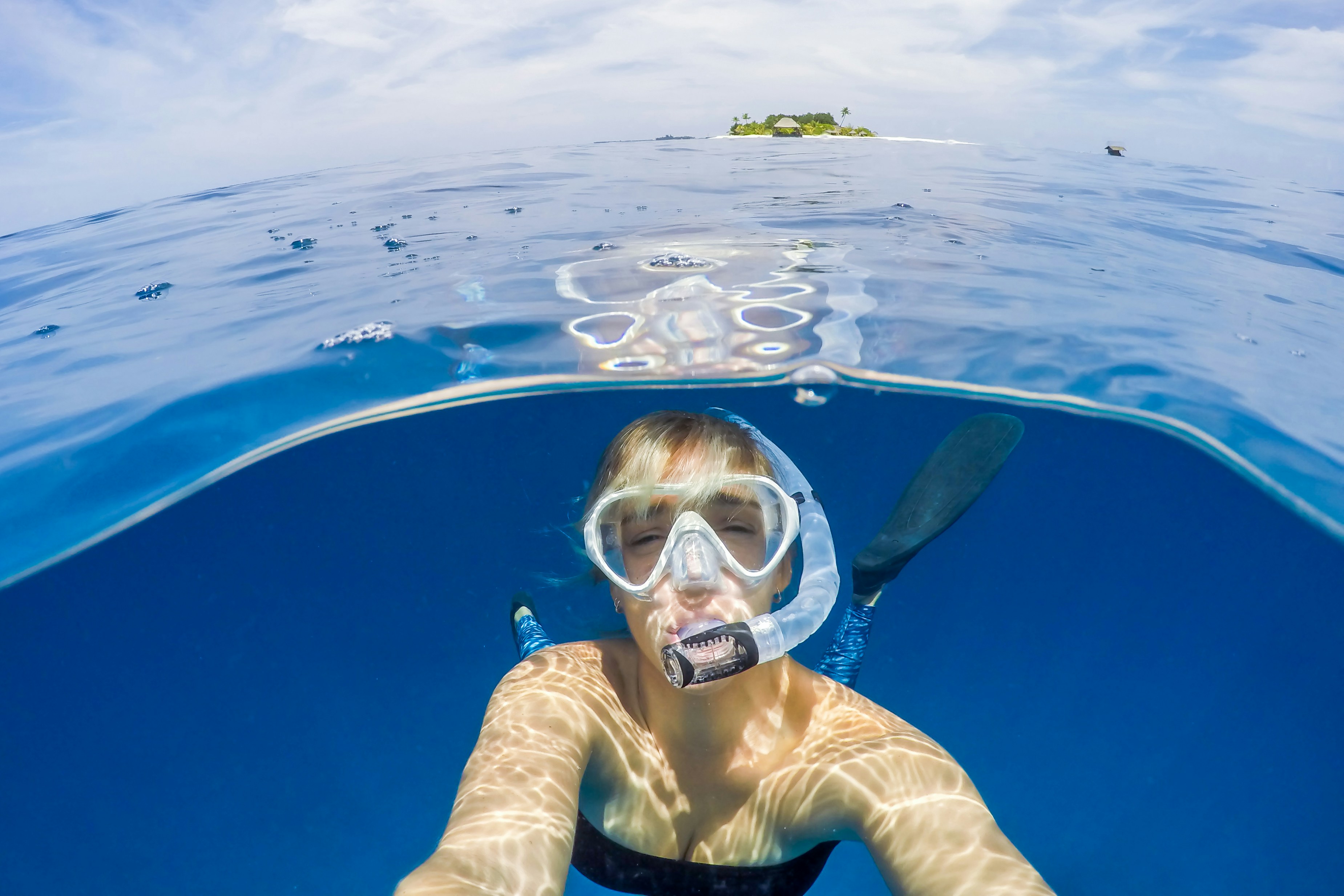 Person snorkeling in clear blue water with a mask and fins, capturing a selfie with a tropical island in the background.