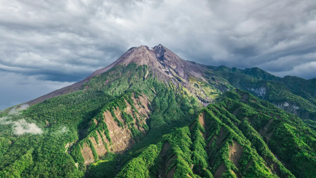 Mount Merapi standing tall against a clear blue sky, with its rugged volcanic slopes visible and the surrounding landscape bathed in sunlight.