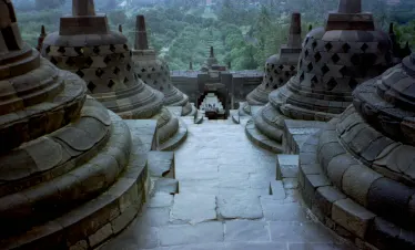 Bird's-eye view of Borobudur Temple, showcasing its tiered structure and intricate stone carvings, leading down to the surrounding green landscape.