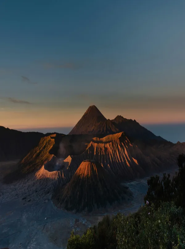 View of Mount Bromo from its base, showcasing the vast plains leading up to the towering volcanic peak, under a clear sky and surrounded by rugged terrain.
